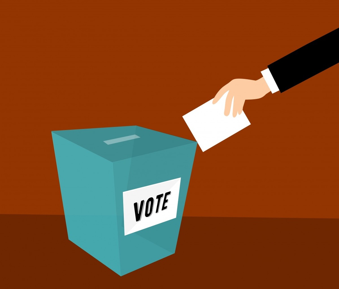 En stiliserad grafisk illustration som visar en hand i kostymärm som lägger en valsedel i en blå röstlåda märkt 'VOTE'. Bilden har en varm orange bakgrund som ger stark kontrast.